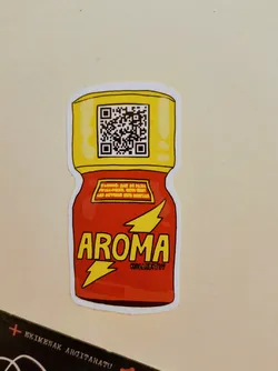 A die-cut sticker shaped like a red bottle with a yellow cap, reminiscent of a hot sauce or popper bottle. The main body is red with the word 'AROMA' in large yellow capital letters, outlined in black. Below 'AROMA' and slightly to the right, in smaller yellow text, is 'KOLLEKTIV'. A yellow lightning bolt symbol is positioned to the left of 'AROMA'. Above 'AROMA', on a yellow rectangular label with a red border, is small, hard-to-read text that appears to be a warning label. The yellow cap features a prominent black and white QR code at its center. The sticker has a clean white border around its entire perimeter.