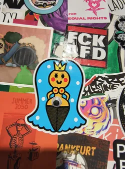 Street sticker The central sticker features a stylized cartoon mermaid with a bright yellow face, rosy cheeks, and black eyes, wearing a golden crown. Her body transitions into a black and yellow patterned skirt, reminiscent of a fishtail. She has large, blue hair or headdress adorned with white polka dots and a googly eye placed centrally on her torso. The sticker has a white border and appears to be die-cut.