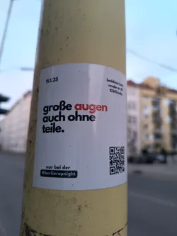 Street sticker A white rectangular sticker with black and red text, adhered to a pole. The sticker reads "große augen auch ohne teile." with "augen" highlighted in red. Below this, it states "nur bei der @berlinrapnight". A QR code is located on the right side of the sticker. The sticker promotes Berlin Rap Night and includes their Instagram handle.