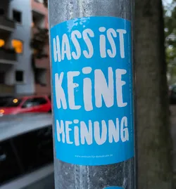 Street sticker The sticker is bright blue with bold, white, slightly irregular sans-serif lettering. The text is arranged in three lines. The top line reads 'HASS IST', the middle line reads 'KEINE', and the bottom line reads 'MEINUNG'. To the right of the main text, in a smaller font, is the text 'Bitte nicht wild verkleben! | V.i.S.d.P.: B. Hannemann, Hasselwerderstraße 38-40, 12439 Berlin'. At the very bottom of the sticker, centered, is a website address: 'www.zentrum-für-demokratie.de'. The sticker appears to be adhered to a metal pole.