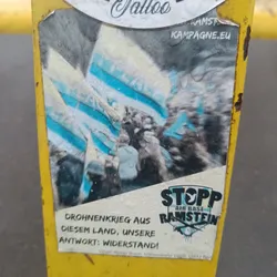 Street sticker A sticker protesting against the Ramstein Air Base's role in the drone war. The sticker features an image of a protest and the text "STOPP AIR BASE RAMSTEIN" in bold letters.  German text translates to "Drone war from this country, our answer: resistance!"