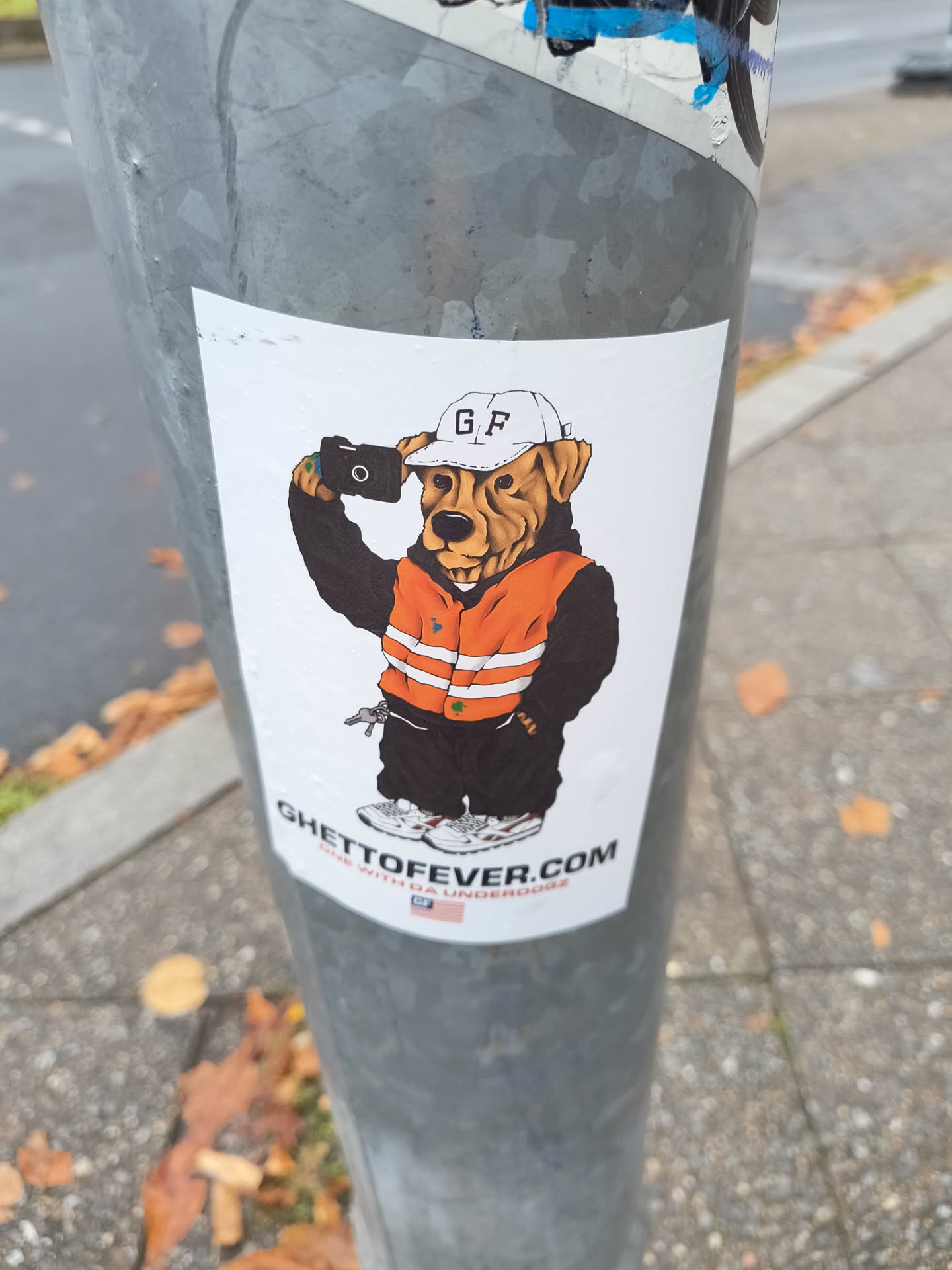 The sticker features a cartoonish dog wearing a white baseball cap with 'GF' embroidered on it, an orange safety vest with reflective stripes over a black hoodie, black pants, and white sneakers. The dog is holding a black camera up to its face as if taking a selfie. The overall style is illustrative and somewhat urban.