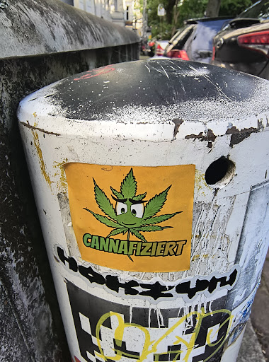 A rectangular yellow sticker featuring a cartoon cannabis leaf with a smiling face and the word 'CANNAFIZIERT' is attached to a white and black bollard on a city street. The sticker is colorful and stands out against the aged bollard.