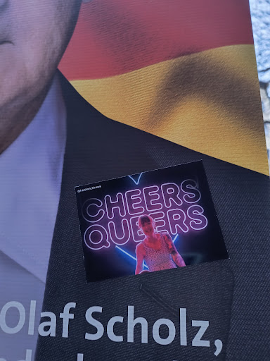 Street sticker A sticker featuring the text 'Cheers Queers' in neon style with an image of a person. The sticker is placed on a dark surface with the text 'Olaf Scholz' visible in the background.