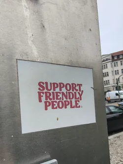The sticker is rectangular with a clean white background. It features the phrase "SUPPORT FRIENDLY PEOPLE" in a bold, rounded, sans-serif font, colored in a bright reddish-pink or coral hue. A small registered trademark symbol (®) is visible after the word "PEOPLE". The sticker appears to be applied to a brushed metallic surface in an outdoor urban environment.