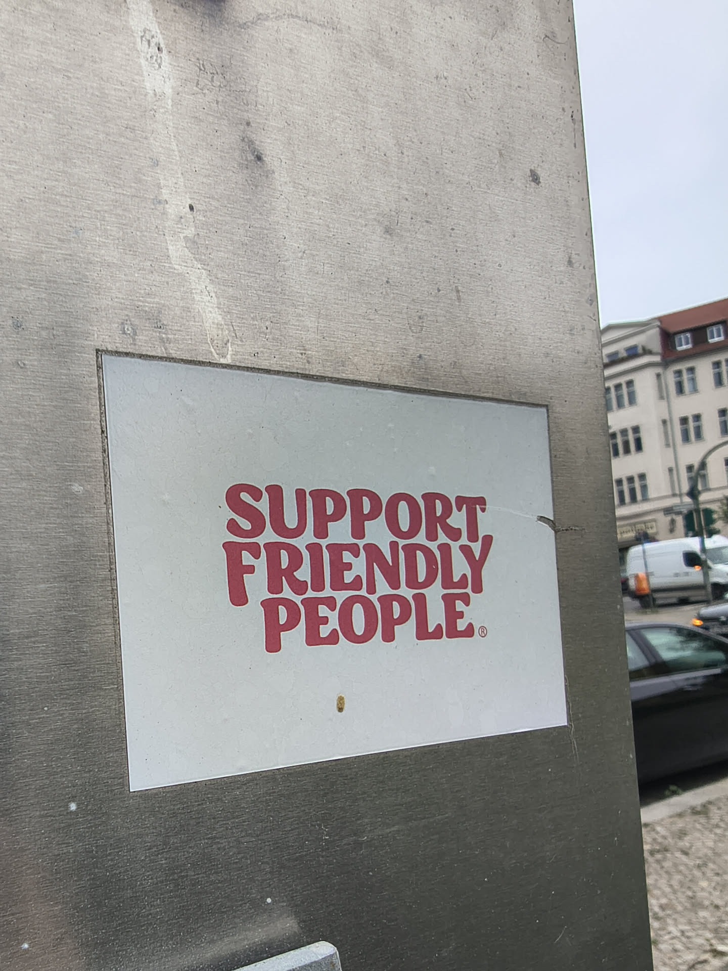 The sticker is rectangular with a clean white background. It features the phrase "SUPPORT FRIENDLY PEOPLE" in a bold, rounded, sans-serif font, colored in a bright reddish-pink or coral hue. A small registered trademark symbol (®) is visible after the word "PEOPLE". The sticker appears to be applied to a brushed metallic surface in an outdoor urban environment.