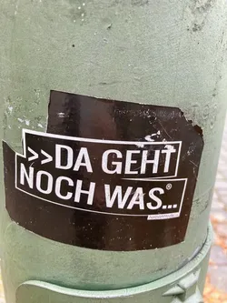 A black sticker with white text on a green pole. The text says: ">Da geht noch was..." which translates to "There's more to come..."