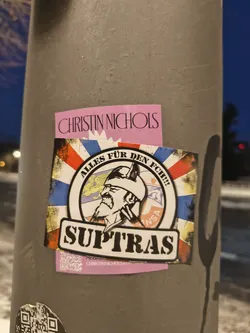 Street sticker The central sticker features a circular emblem set against a distressed, grunge-style background resembling a flag with red, white, blue, and yellow sections. The emblem depicts a profile of a bearded man wearing a horned helmet, reminiscent of a Viking warrior. Above his head within the circle is a yellow eagle. Around the top arc of the circle, the text "ALLES FÜR DEN FCH!!!" is written. A smaller text "INSA" is visible on the right side of the circle. Below the circular emblem, a black banner with a white outline prominently displays the word "SUPTRAS". The overall aesthetic is rugged and fan-oriented.