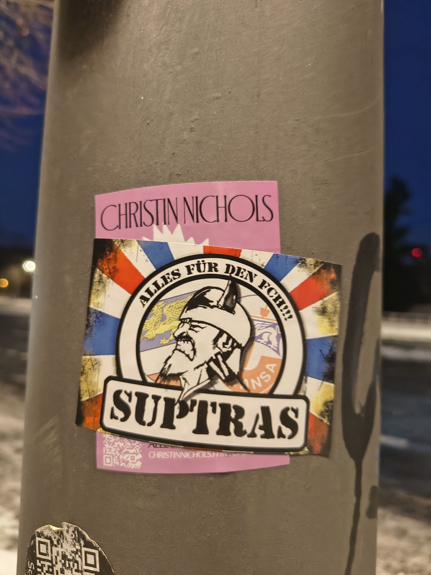 The central sticker features a circular emblem set against a distressed, grunge-style background resembling a flag with red, white, blue, and yellow sections. The emblem depicts a profile of a bearded man wearing a horned helmet, reminiscent of a Viking warrior. Above his head within the circle is a yellow eagle. Around the top arc of the circle, the text "ALLES FÜR DEN FCH!!!" is written. A smaller text "INSA" is visible on the right side of the circle. Below the circular emblem, a black banner with a white outline prominently displays the word "SUPTRAS". The overall aesthetic is rugged and fan-oriented.