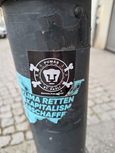 Street sticker This is a black sticker with the words PUMAS ST. PAULI in white text. The sticker also features an image of a stylized anchor and bones, along with the years 2017 and 2020. The sticker is located on a pole.