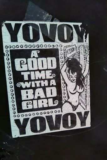 Street sticker A rectangular sticker featuring a black and white design. The sticker has a cracked and aged appearance, suggesting it has been in place for some time. The design includes a drawing of a woman, and bold lettering that reads "A GOOD TIME WITH A BAD GIRL". The top and bottom of the design are framed by the word "Yovoy".