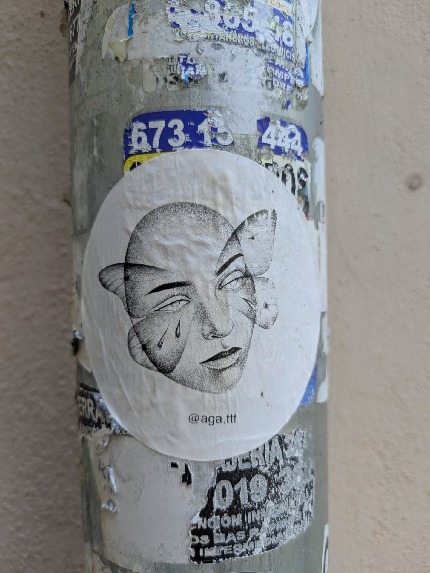 This round sticker is adhered to a textured, weathered pole covered in layers of other stickers and peeling posters. The sticker's design is a stylized black and white drawing of a face with butterfly wings in place of some features. The face appears to have one tear, and there is text at the bottom of the sticker.
