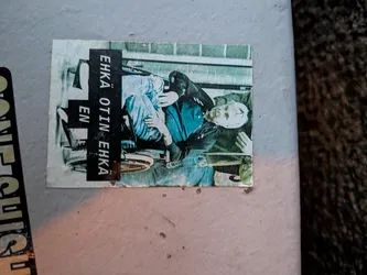 The sticker is rectangular and appears to be made of paper. It depicts a person, likely an elderly man, seated in a wheelchair. The person is wearing a dark jacket and light blue pants. The background is a muted green, possibly representing a wall or a building. There is a black vertical band on the left side of the sticker with white text. The overall color palette is desaturated, giving it a vintage or aged appearance. Some parts of the sticker appear worn or torn.