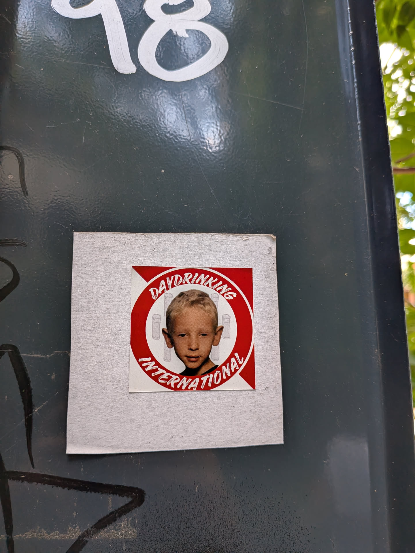Square sticker with a central red circular emblem. Inside is a photo of a young boy against a red and white striped background with faint beer glass icons. The text 'DAYDRINKING INTERNATIONAL' wraps around the circle.