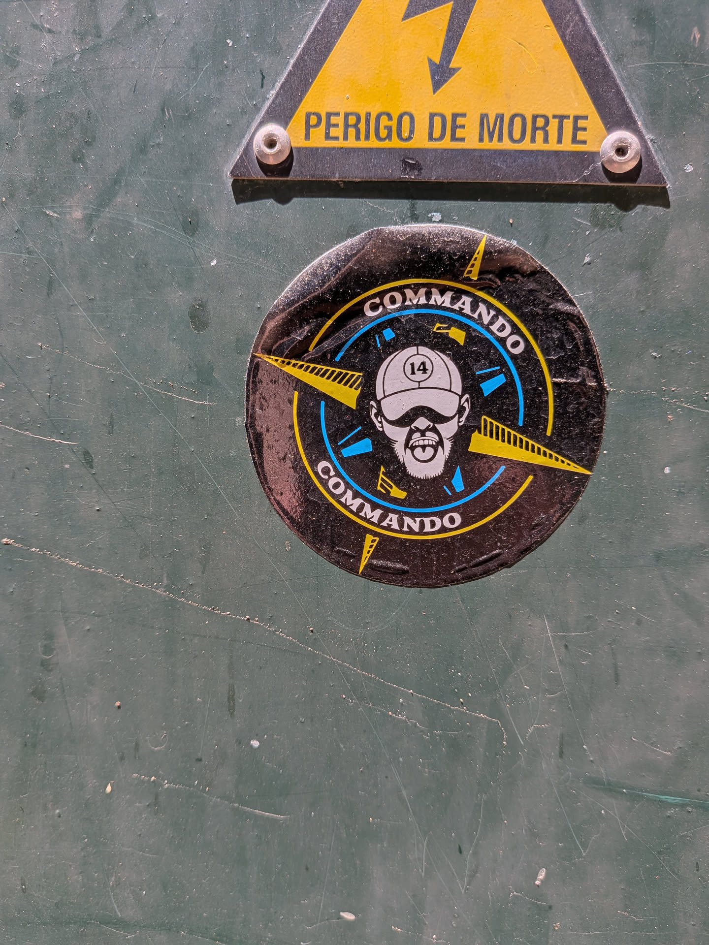 Circular black sticker depicting a bearded man in a cap (marked '14') and sunglasses. The portrait is enclosed by blue and yellow circular compass motifs, with 'COMMANDO' printed in white at the top and bottom.