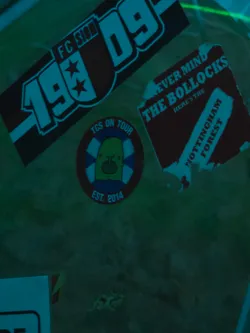 Street sticker The image features three distinct stickers on a dark, possibly textured, surface. The top-left sticker is a rectangular design for 'FC SION 1909', presented with a dark red background, white outlines, and bold white text, including two stars. Below and slightly to the right, a circular sticker displays a light green, blob-like character with large eyes and a small mouth, holding a red object, set against a blue and white striped background resembling a lifebuoy. This circular sticker is framed by a dark red outer ring with white text reading 'TCS ON TOUR EST. 2014'. To the top-right, a roughly rectangular sticker with torn edges features a dark red background and prominent white text that reads 'NEVER MIND THE BOLLOCKS HERE'S THE NOTTINGHAM FOREST', a clear reference to the iconic Sex Pistols album.