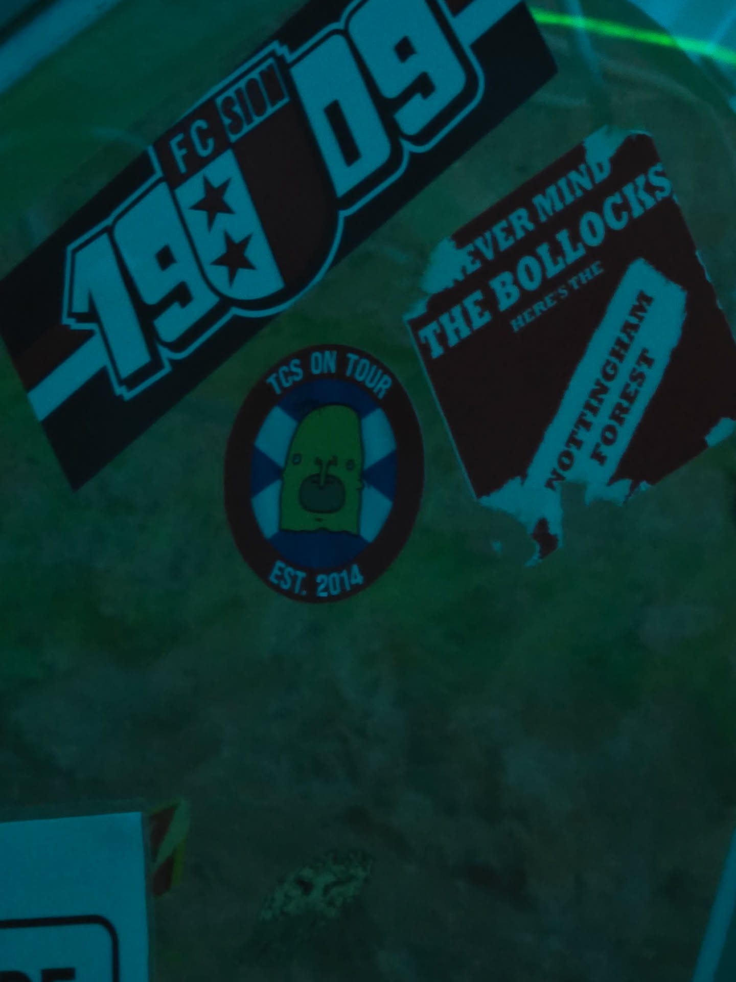 The image features three distinct stickers on a dark, possibly textured, surface. The top-left sticker is a rectangular design for 'FC SION 1909', presented with a dark red background, white outlines, and bold white text, including two stars. Below and slightly to the right, a circular sticker displays a light green, blob-like character with large eyes and a small mouth, holding a red object, set against a blue and white striped background resembling a lifebuoy. This circular sticker is framed by a dark red outer ring with white text reading 'TCS ON TOUR EST. 2014'. To the top-right, a roughly rectangular sticker with torn edges features a dark red background and prominent white text that reads 'NEVER MIND THE BOLLOCKS HERE'S THE NOTTINGHAM FOREST', a clear reference to the iconic Sex Pistols album.