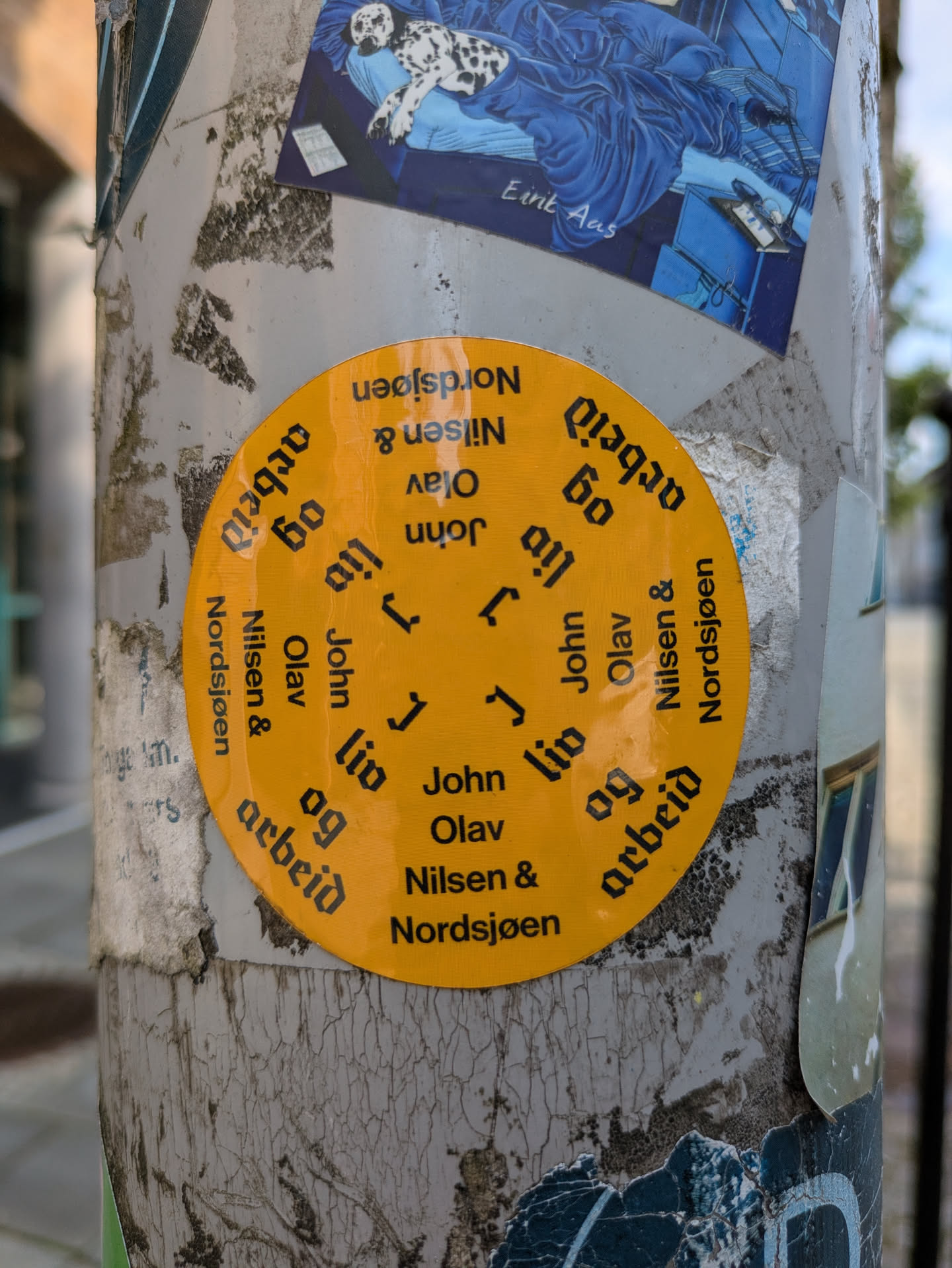 The central object is a large, bright yellow circular sticker with black text arranged radially. The text reads "John Olav Nilsen & Nordsjøen" and "liv og arbeid", repeated several times around the circle. The sticker is placed on a light grey pole that shows signs of wear, peeling paint, and remnants of other old stickers.