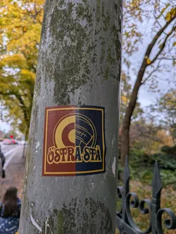 Street sticker The sticker is square with a yellow border. The left half of the sticker is a deep red color, featuring a large yellow 'O' with a smaller yellow circle inside it. The right half of the sticker is a dark blue color, depicting a stylized football with visible stitching lines. Below the emblem, the text 'ÖSTRÅSTÅ' is written in a bold, decorative yellow font with a red outline.