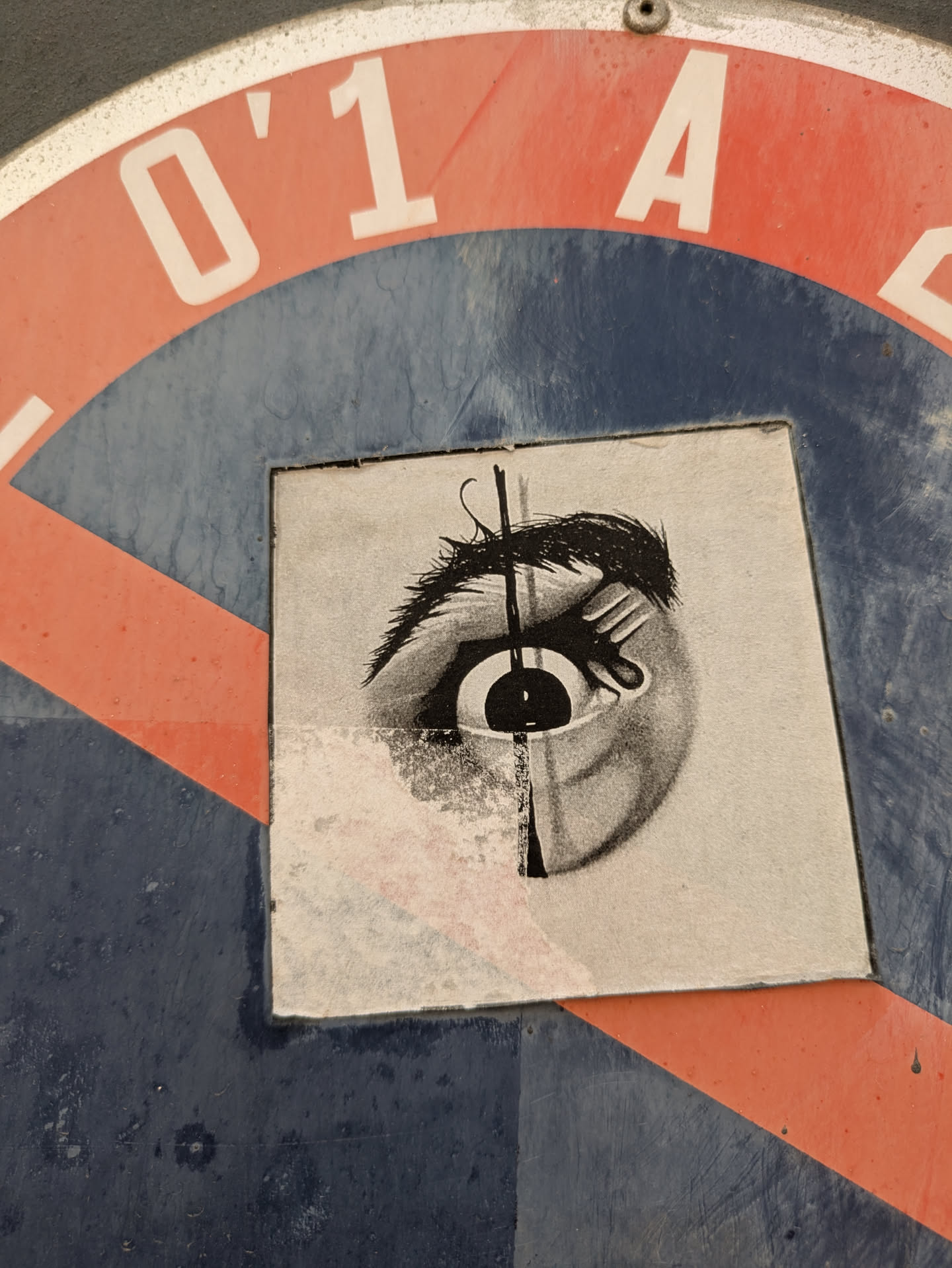 A square black and white sticker on a blue and red surface depicts an eye with a line running through it. The sticker is slightly damaged at the bottom.