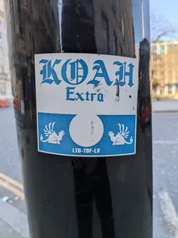Street sticker A rectangular sticker with a white background on the top half and a light blue background on the bottom half. The top features the word "KOAH" in a large, ornate, gothic-style blue font, with "Extra" in a smaller, simpler blue serif font beneath it. The bottom half, set against a light blue background, displays two stylized white winged creatures (resembling winged camels or dromedaries) facing inwards towards a central white circle. Below these figures, the text "LTB-TBF-LV" is printed in white. The sticker is applied to a black pole and shows minor signs of wear, including a small tear near the 'H' in KOAH.