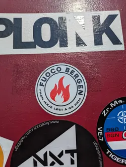 Street sticker The sticker is round with a white background and a thick black border. In the center, there are three stylized red flames. Arched along the top perimeter in black capital letters is 'FUOCO BERGEN', and along the bottom, 'FAEN IKKJE LØST Å GÅ HEM'. The overall design is clean and bold.