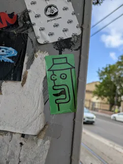 Street sticker The sticker features a simple, cartoon-like face drawn with black lines on a green background. The face has large, round eyes and a prominent mouth. A hat is also drawn on top of the head. The sticker has the name "semo" written on the right side of it. It is adhered to a grey, weathered street pole, surrounded by other stickers and peeling posters.