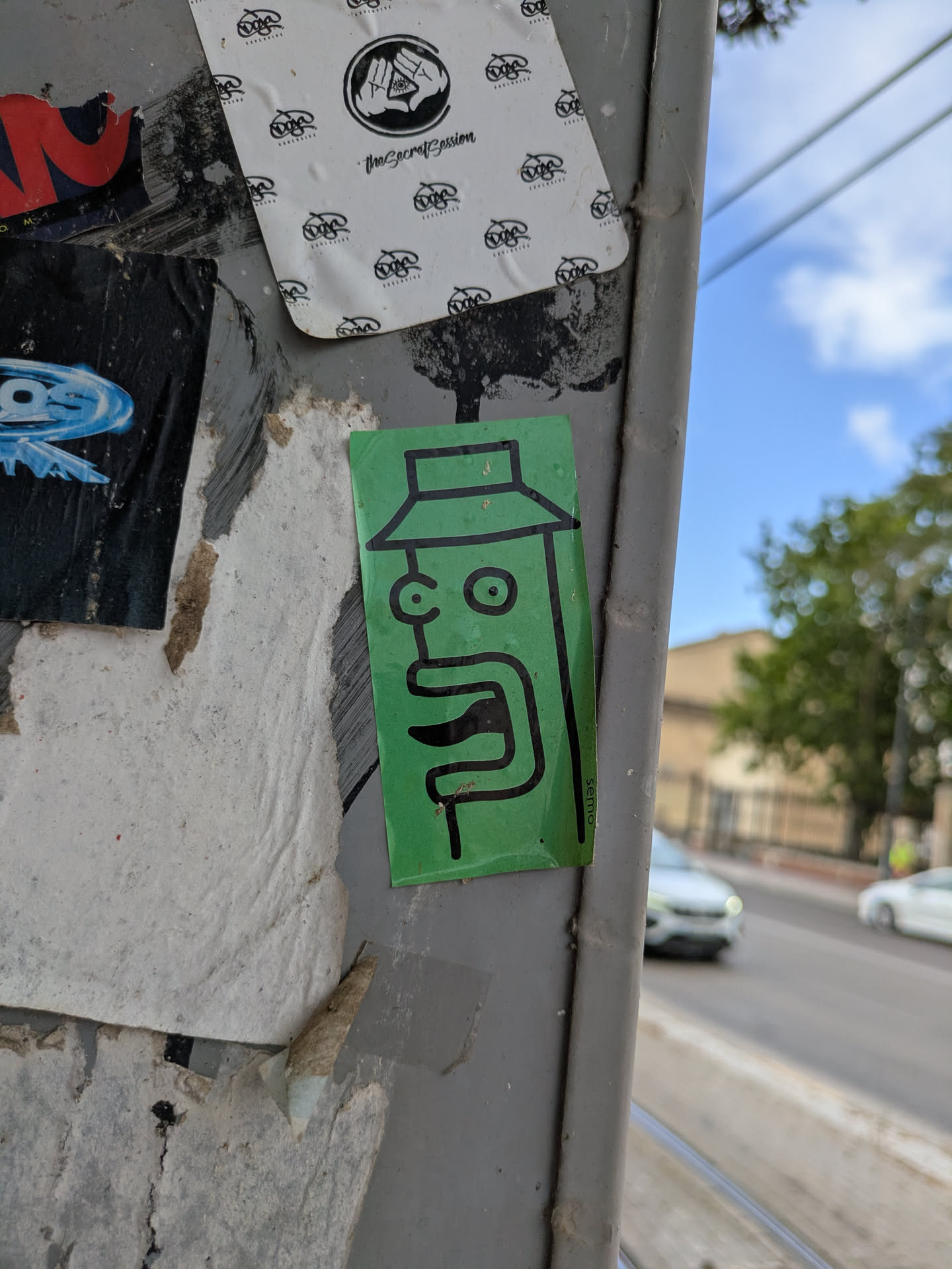 Street sticker The sticker features a simple, cartoon-like face drawn with black lines on a green background. The face has large, round eyes and a prominent mouth. A hat is also drawn on top of the head. The sticker has the name "semo" written on the right side of it. It is adhered to a grey, weathered street pole, surrounded by other stickers and peeling posters.