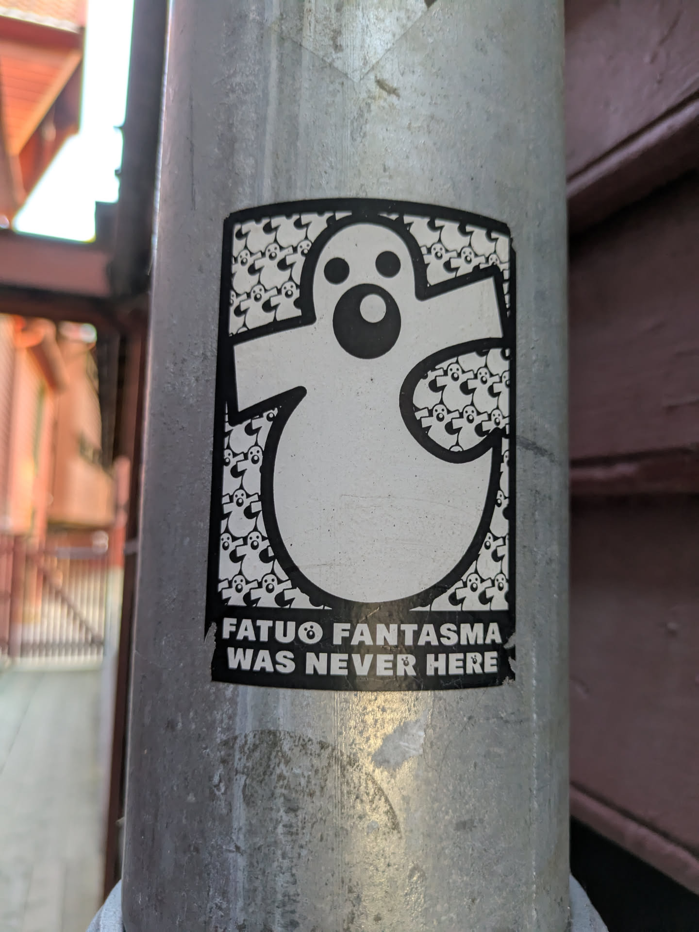 This sticker showcases a prominent black-outlined white ghost figure with simple, circular eyes and mouth. The interior of the large ghost is intricately filled with a repeating pattern of much smaller, identical ghost characters, creating a unique texture. Below the main graphic, white capitalized text on a black background reads 'FATUO FANTASMA' followed by a small ghost icon, and then 'WAS NEVER HERE'. The overall design is monochrome and features a playful yet slightly mysterious aesthetic. The sticker is affixed to a metallic pole.