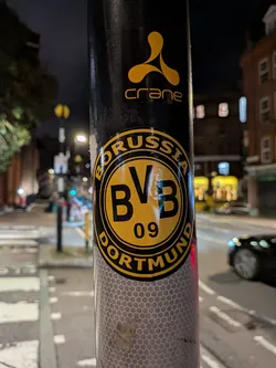 Street sticker A circular sticker featuring the emblem of the German football club Borussia Dortmund. The sticker is predominantly yellow and black. It has a thick black outer ring with the word "BORUSSIA" in yellow capital letters at the top and "DORTMUND" in yellow capital letters at the bottom. The inner circle is bright yellow with a thick black border, containing the large black capital letters "BVB" stacked vertically, and below them, the number "09" in black. The sticker shows some minor wear and tear, indicating it's a physical sticker applied to a black pole.