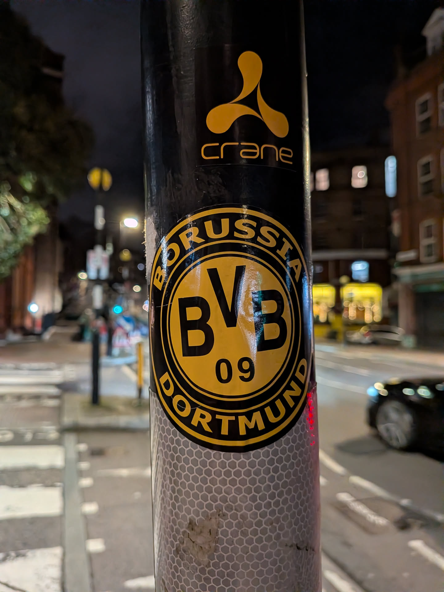A circular sticker featuring the emblem of the German football club Borussia Dortmund. The sticker is predominantly yellow and black. It has a thick black outer ring with the word "BORUSSIA" in yellow capital letters at the top and "DORTMUND" in yellow capital letters at the bottom. The inner circle is bright yellow with a thick black border, containing the large black capital letters "BVB" stacked vertically, and below them, the number "09" in black. The sticker shows some minor wear and tear, indicating it's a physical sticker applied to a black pole.