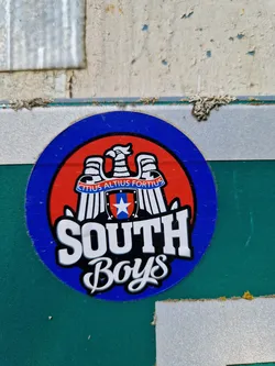 Street sticker A round sticker featuring a red, white, and blue design.  The center depicts a heraldic eagle above a shield with a star, surrounded by a banner with the text "Citius Altius Fortius." Below the emblem, it says "SOUTH Boys" in stylized lettering. The sticker has a blue outer ring and is affixed to a teal surface.
