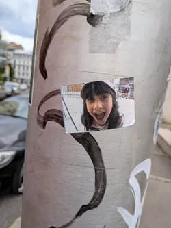 Street sticker A rectangular photo sticker on a metal pole showing a young girl with dark bangs and a hood, smiling widely with an open mouth. The background is a slightly blurred outdoor scene.