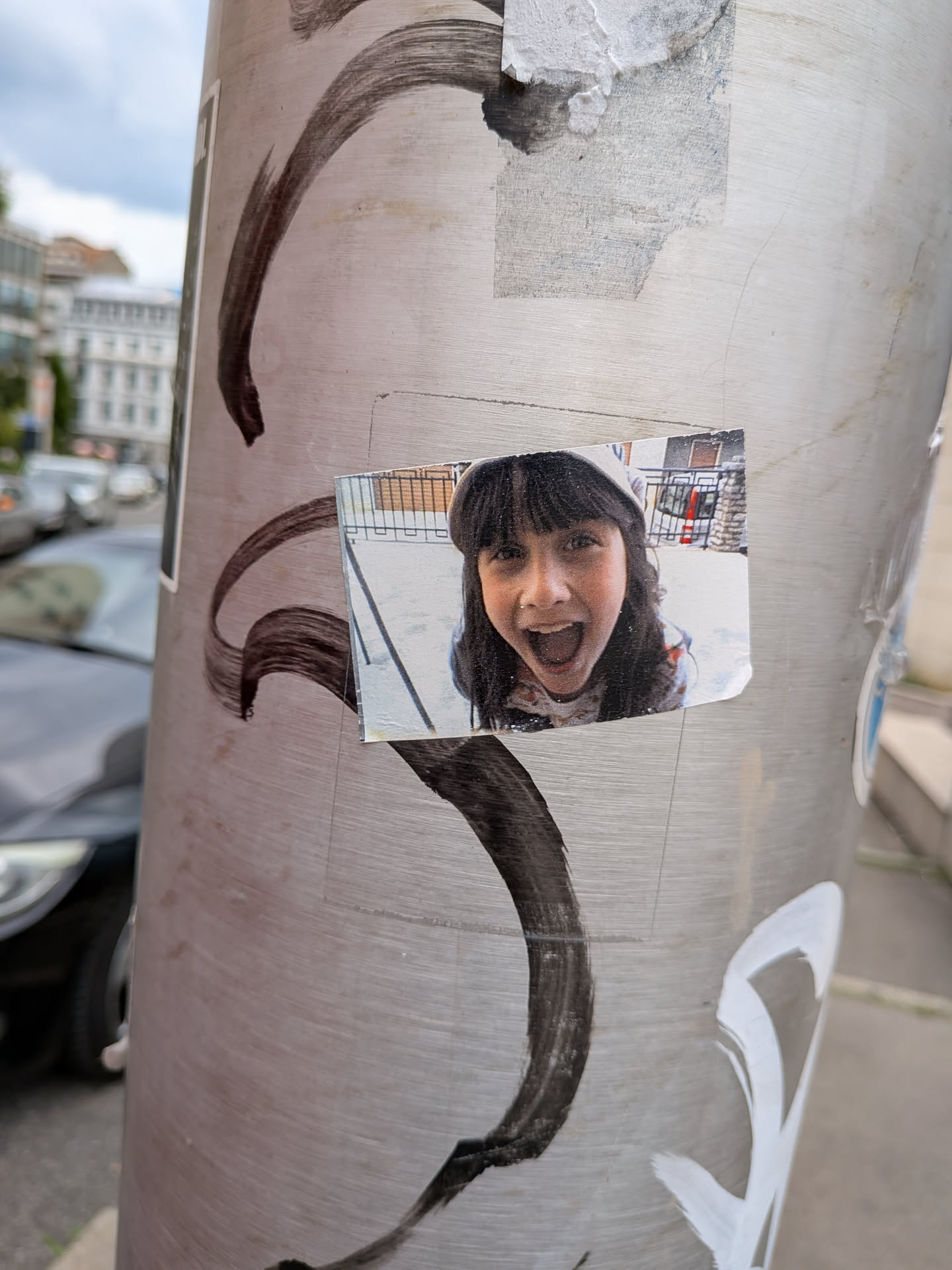 A rectangular photo sticker on a metal pole showing a young girl with dark bangs and a hood, smiling widely with an open mouth. The background is a slightly blurred outdoor scene.