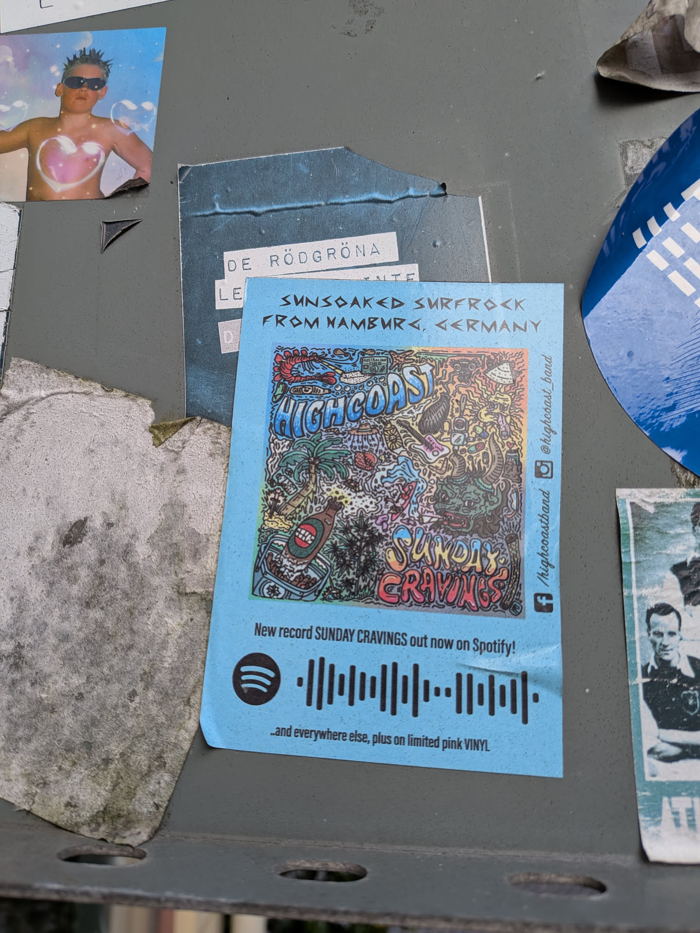 The sticker is predominantly light blue and features a busy, colorful, and somewhat psychedelic illustration. At the top, the words 'HIGHCOAST' are prominently displayed in large, stylized blue lettering. Below this, the text 'SUNDAY CRAVINGS' is written in a bubbly, multi-colored font. The illustration itself is dense with various elements, including palm trees, a beer bottle in a cooler, a lobster, a monster-like creature, musical instruments (guitar, drums), and abstract patterns. To the right of the illustration, vertically oriented text reads '@highcoast_band'. Below the main illustration, there is text indicating 'New record SUNDAY CRAVINGS out now on Spotify!' followed by a Spotify logo and a Spotify code. At the very bottom, it says '...and everywhere else, plus on limited pink VINYL'. The sticker appears to be slightly creased and worn, suggesting it has been applied for some time.