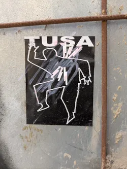 This black sticker features a white outline of a dancing or gesturing figure. The figure is stylized and abstract, with jagged lines and sharp angles. Above the figure, the word TUSA is printed in bold, white letters. The sticker is affixed to a weathered metal surface.