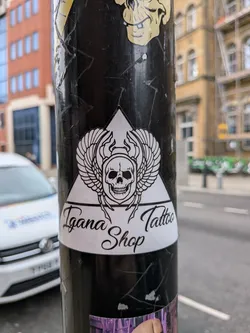 Street sticker The sticker features a bold, black and white design centered around a stylized skull with large, feathered wings extending outwards. The skull has a detailed, somewhat menacing expression with visible teeth and appears to be wearing a horned helmet or crown. This central winged skull motif is set against a white background within a prominent black-outlined triangle. Below the triangle, the words "Igana Tattoo Shop" are written in an elegant, flowing black script on a white banner-like section. The overall aesthetic is clean, graphic, and has a distinct tattoo art style.