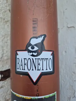 Street sticker A black and white sticker with a stylized illustration of an eye with a mustache and a single tear. Below the illustration is the text "BAFONETTO" in a bold, sans-serif font.  The sticker is applied to a brown pipe.