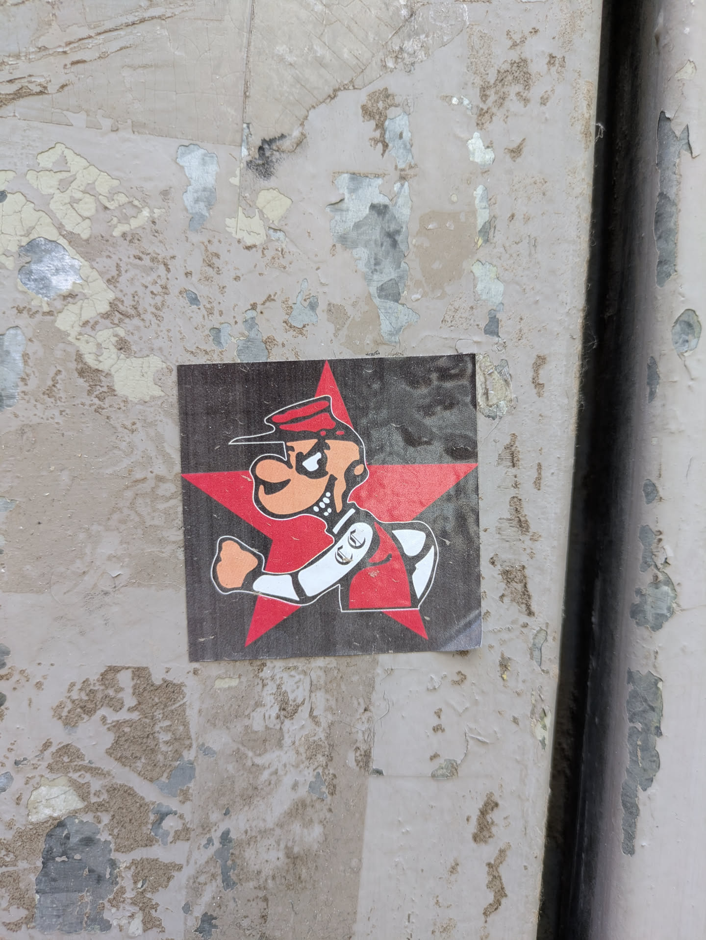 The sticker features a cartoon character with an orange face and a sinister expression, wearing a red cap and uniform with white trim. The character is posed against a bright red star on a dark grey background. The sticker is affixed to a peeling painted surface.
