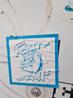 Street sticker A light-blue square sticker featuring a pixelated bulldog graphic in a bold, blue color scheme. The sticker displays a stylized bulldog head with sharp teeth, surrounded by pixelated geometric patterns. Some of the text is partially damaged, but it shows some glitch effect or pixelation. The sticker has a unique, somewhat aggressive aesthetic. 