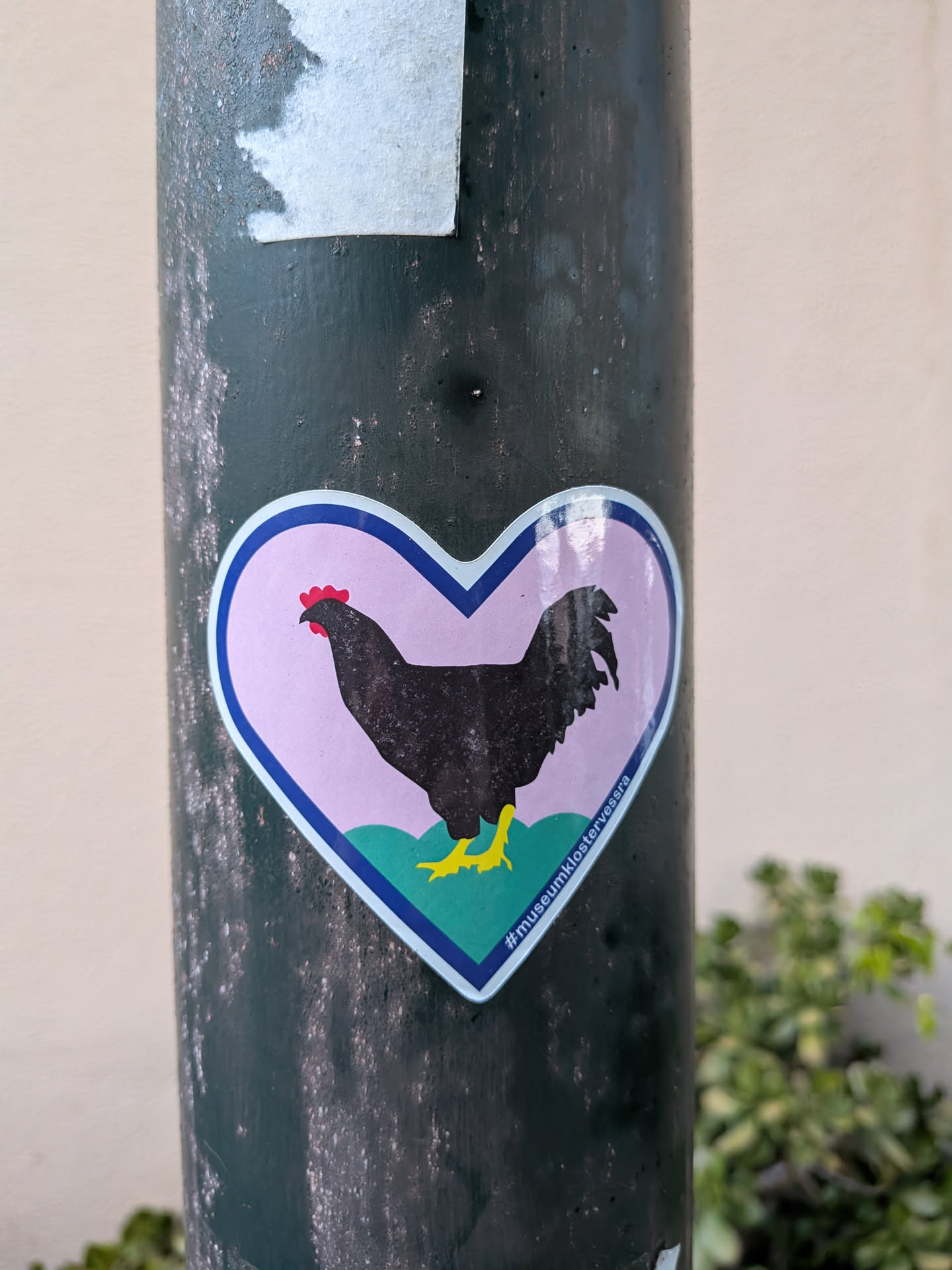A heart-shaped sticker featuring a black chicken silhouette with a red comb and yellow feet, standing on green hills against a light purple background.