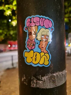 A sticker featuring cartoonish illustrations of two women, one with pink hair and the other with blonde hair, both seemingly topless. The style is reminiscent of graffiti art, with bubble-like lettering forming 'GLSVN' and 'TØP' on the sticker. 