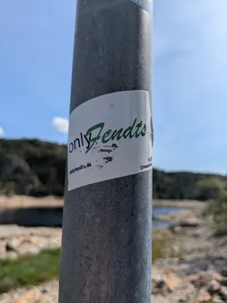 Street sticker A rectangular white sticker is wrapped around a grey metal pole. The dominant text on the sticker reads 'onlyFendts', with 'only' in black and 'Fendts' in a distinctive green font, suggesting a brand or fan community related to Fendt, a well-known agricultural machinery manufacturer. Below this, a website address 'onlyfendts.de' is visible. To the right, partially obscured, an Instagram icon and the handle '@klaugsahna' are present. The sticker appears slightly worn or peeled in the middle, revealing some underlying damage. The background shows a natural landscape with a body of water and distant hills under a clear blue sky, implying an outdoor setting.