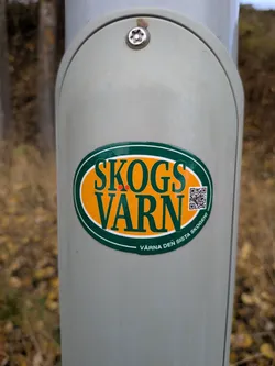 Street sticker The sticker is oval-shaped with a dark green border. Inside the border, the background is a bright orange. The text 'SKOGS VÄRN' is prominently displayed in large, dark green, sans-serif capital letters. Two small red dots are positioned above the 'V' in 'VÄRN'. To the right of the text, a black and white QR code is visible. Along the bottom curve of the sticker, in smaller dark green text, it reads 'VÄRNA DEN SISTA SKOGEN!'. The sticker appears to be adhered to a light gray, cylindrical object.