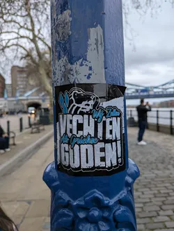 Street sticker A rectangular sticker predominantly black, featuring a stylized, fierce animal head (resembling a lion or bear) at the top, outlined in white with light blue diamond shapes. Below the animal head, the main text "VECHTEN" is prominently displayed in large, distressed white capital letters with a black outline. Above "VECHTEN", in a smaller, script-like light blue font, are the words "Wij Zullen". Below "VECHTEN", also in script-like light blue font, are "Als Griekse", followed by "GUDEN!" in large, distressed white capital letters with a black outline at the bottom. The sticker has a gritty, impactful aesthetic and is applied to a blue painted pole, showing slight wear and curling at the edges.