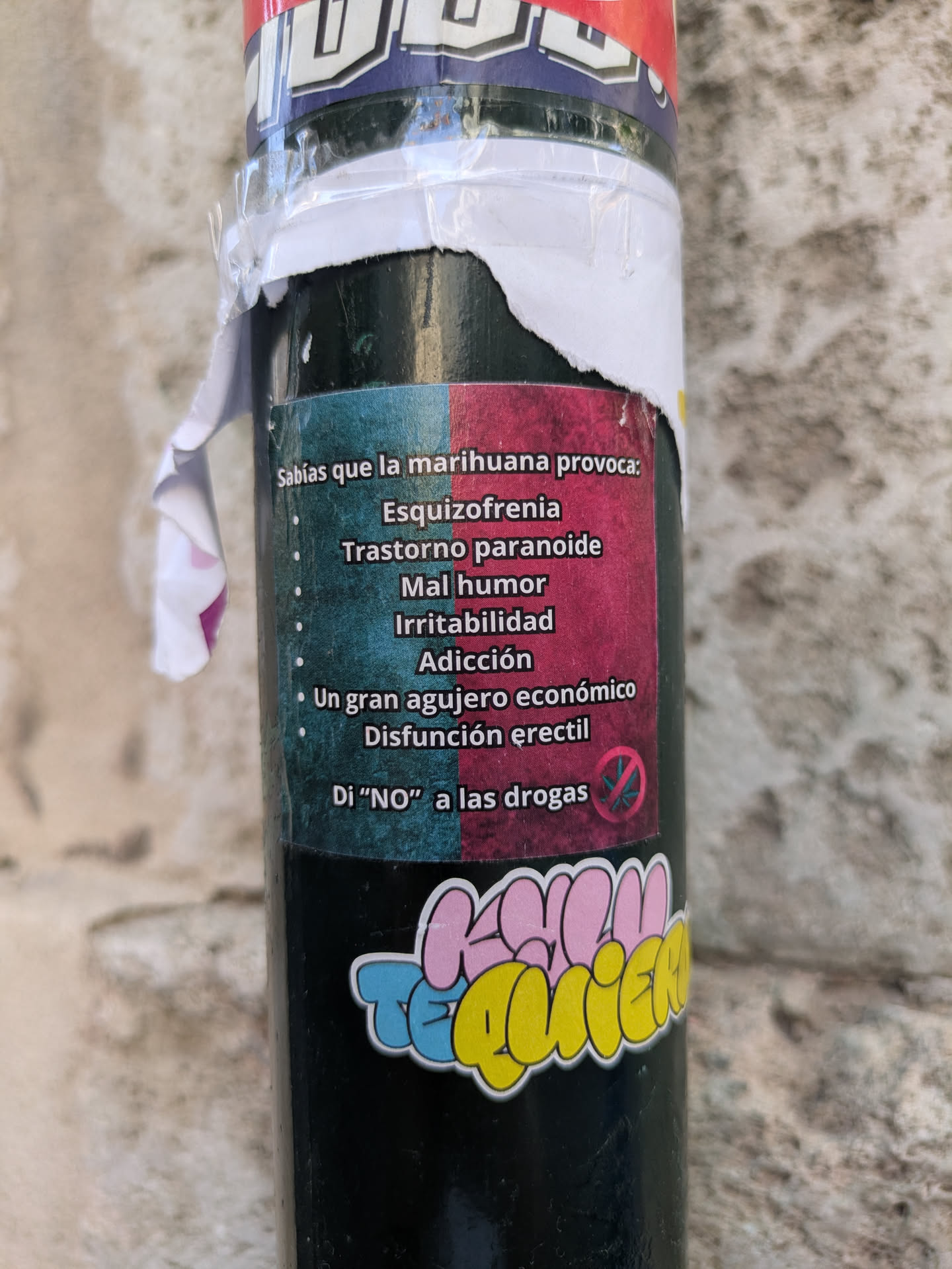 The image features a pole covered with stickers. The primary sticker in focus is an anti-marijuana message listing negative health and economic consequences of marijuana use in Spanish. Below it, there is a colorful sticker with text.