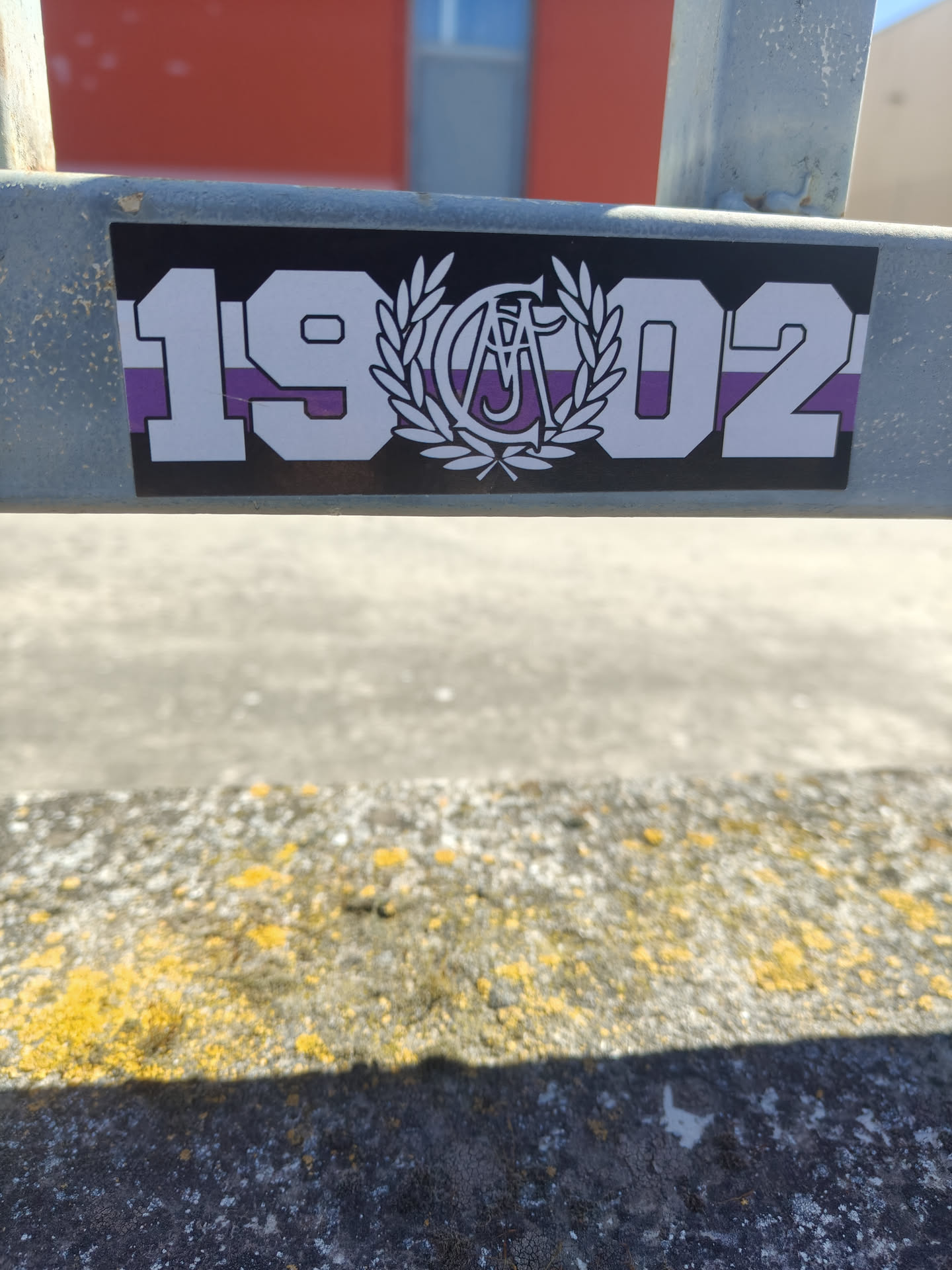 A rectangular sticker with a black border, featuring a design of bold white numbers '19' on the left and '02' on the right. In the center, between the numbers, is a white emblem consisting of a laurel wreath surrounding intertwined letters, which appear to be 'C' and 'A' or 'A' and 'C'. The background of the sticker, including the fill of the central emblem's letters, is horizontally striped in alternating purple and white bands. The sticker is adhered to a grey metal beam.