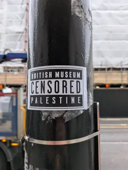Street sticker A rectangular, black and white sticker affixed to a textured black pole. The sticker features a black background with a thin white border. It contains three lines of white, sans-serif, uppercase text. The top line reads 'BRITISH MUSEUM', the middle line, which is significantly larger and bolder, reads 'CENSORED', and the bottom line reads 'PALESTINE'. The sticker shows minor signs of wear, with slight peeling visible at the edges.