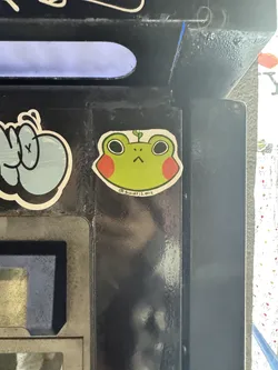 This is a die-cut sticker featuring a cartoon-style frog head. The frog is primarily light green with large black, circular eyes and a small, downturned V-shaped mouth, giving it a somewhat grumpy or unamused expression. It has distinct round, reddish-orange blush marks on its cheeks. A small green sprout or leaf is growing from the top of its head. The entire design is outlined in white, and below the frog, there's small black text.