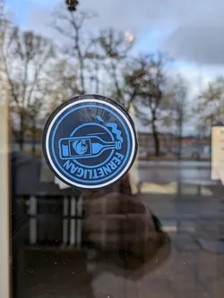 Street sticker The sticker is circular with a black outer ring and a white inner ring. Inside, the background is black. A blue outline depicts a bottle with a monkey's head visible inside. Blue liquid is splashing out of the bottle's neck. The text 'FERNETLIGAN' is written in blue, curved lettering around the bottom half of the sticker.